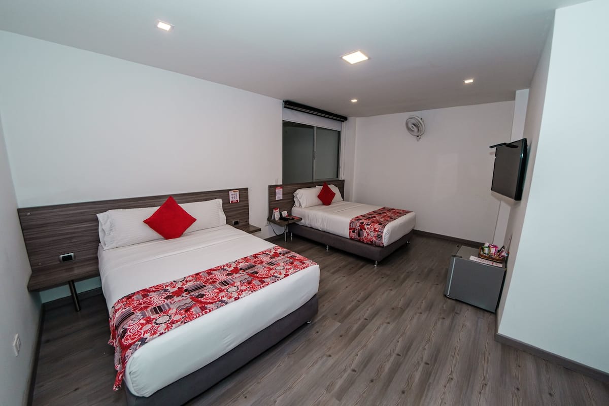 A hotel room with two beds, each with a red pillow, a bedside table, and a small desk with a chair.