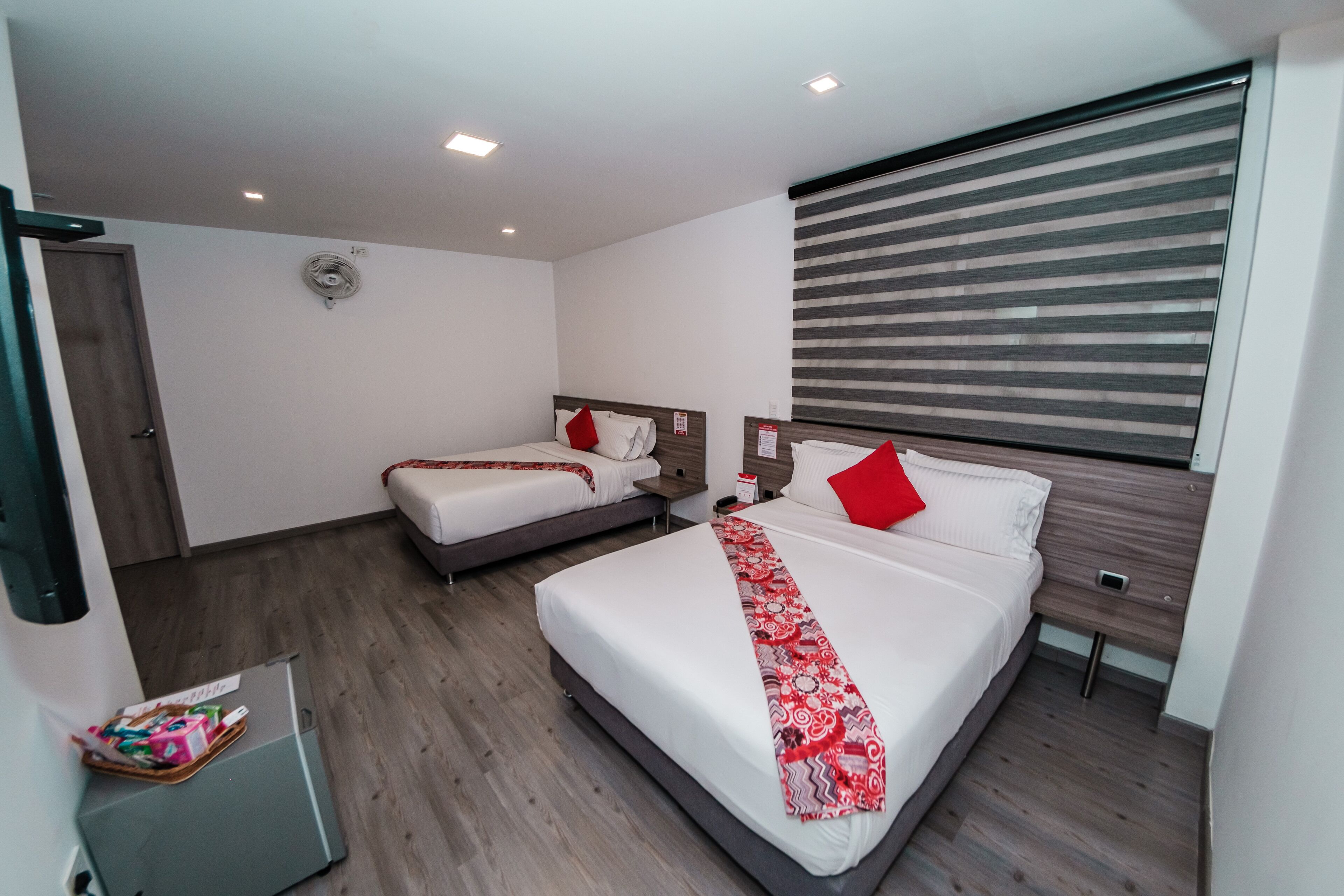 Une chambre d’hôtel moderne avec deux lits, un plancher en bois, une grande fenêtre avec stores et un petit bureau doté d’une lampe.