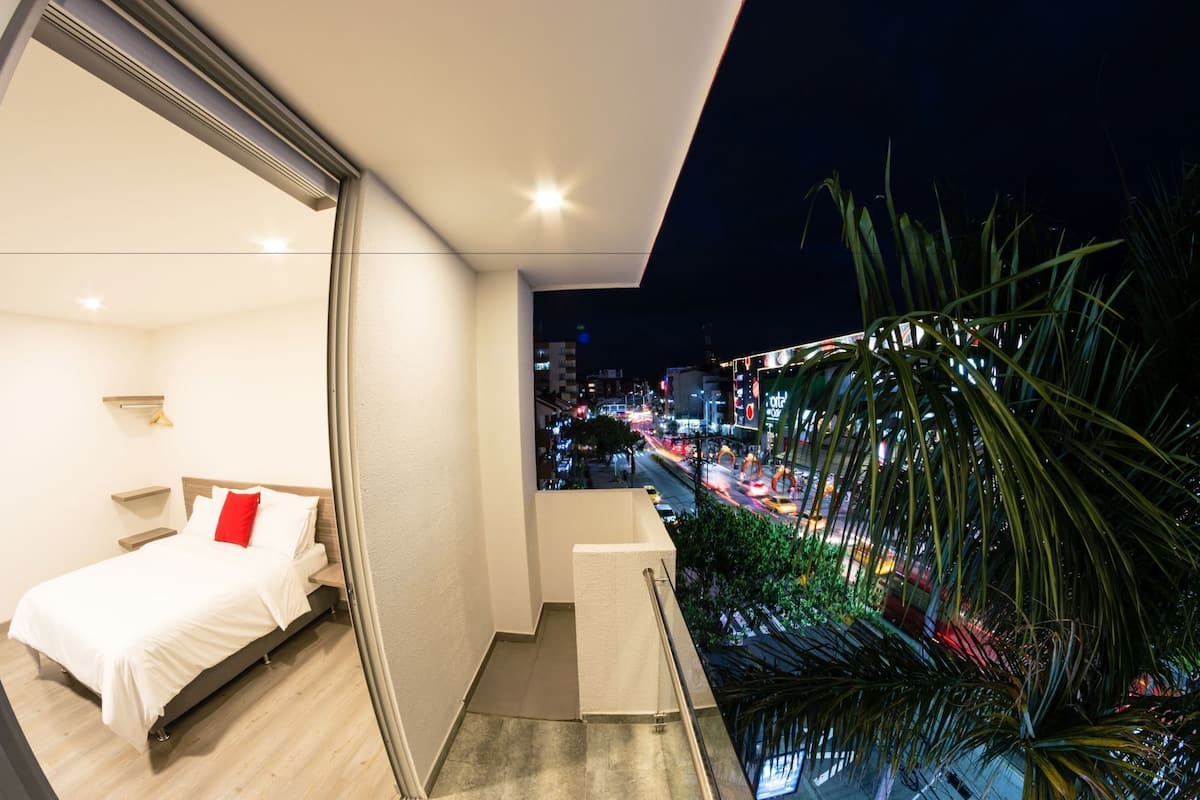 A modern hotel room with a bed, a small seating area, and a balcony with a city view.