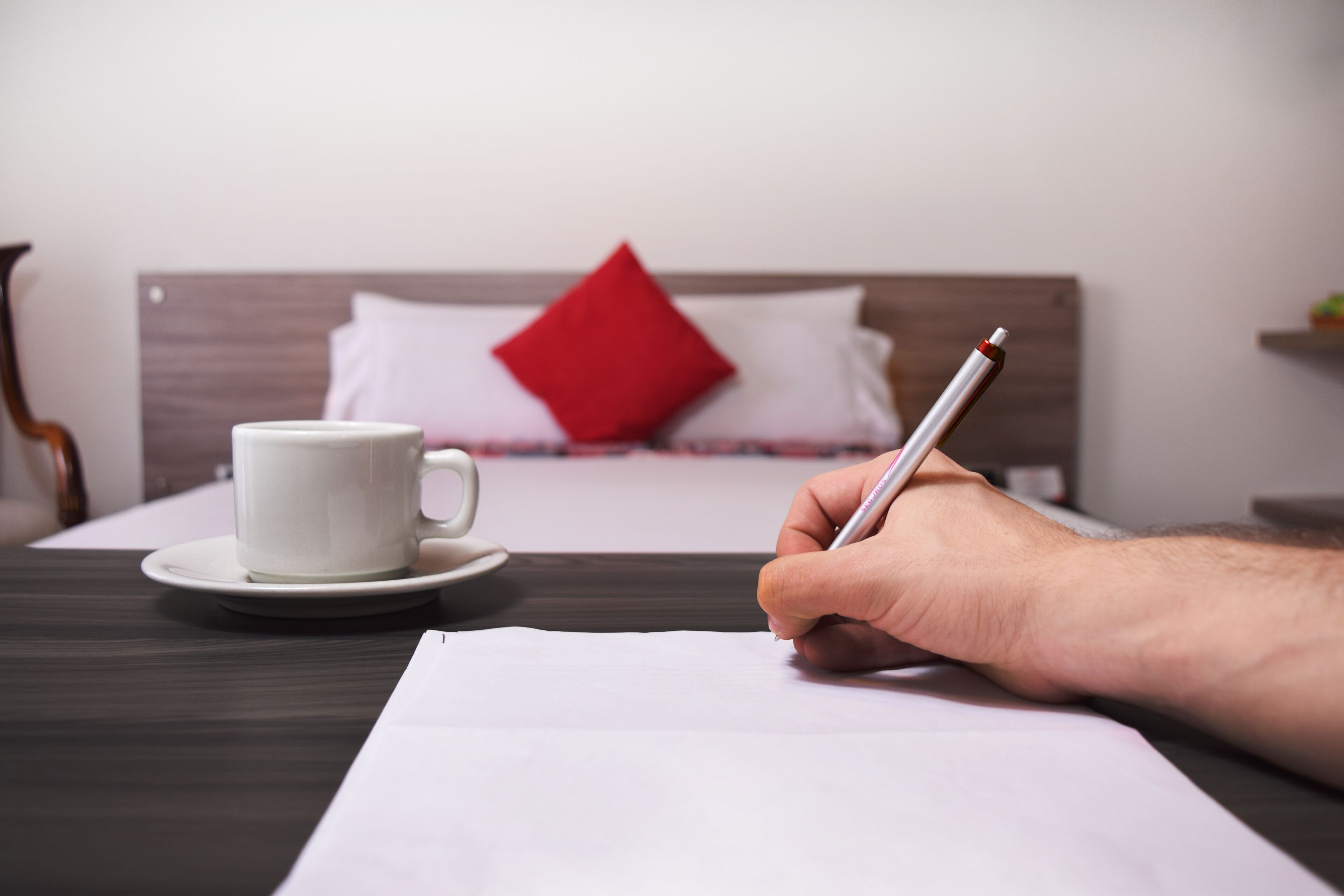 A hand holding a pen, writing on a piece of paper, with a white cup and saucer on a table in front of a bed with a red pillow.