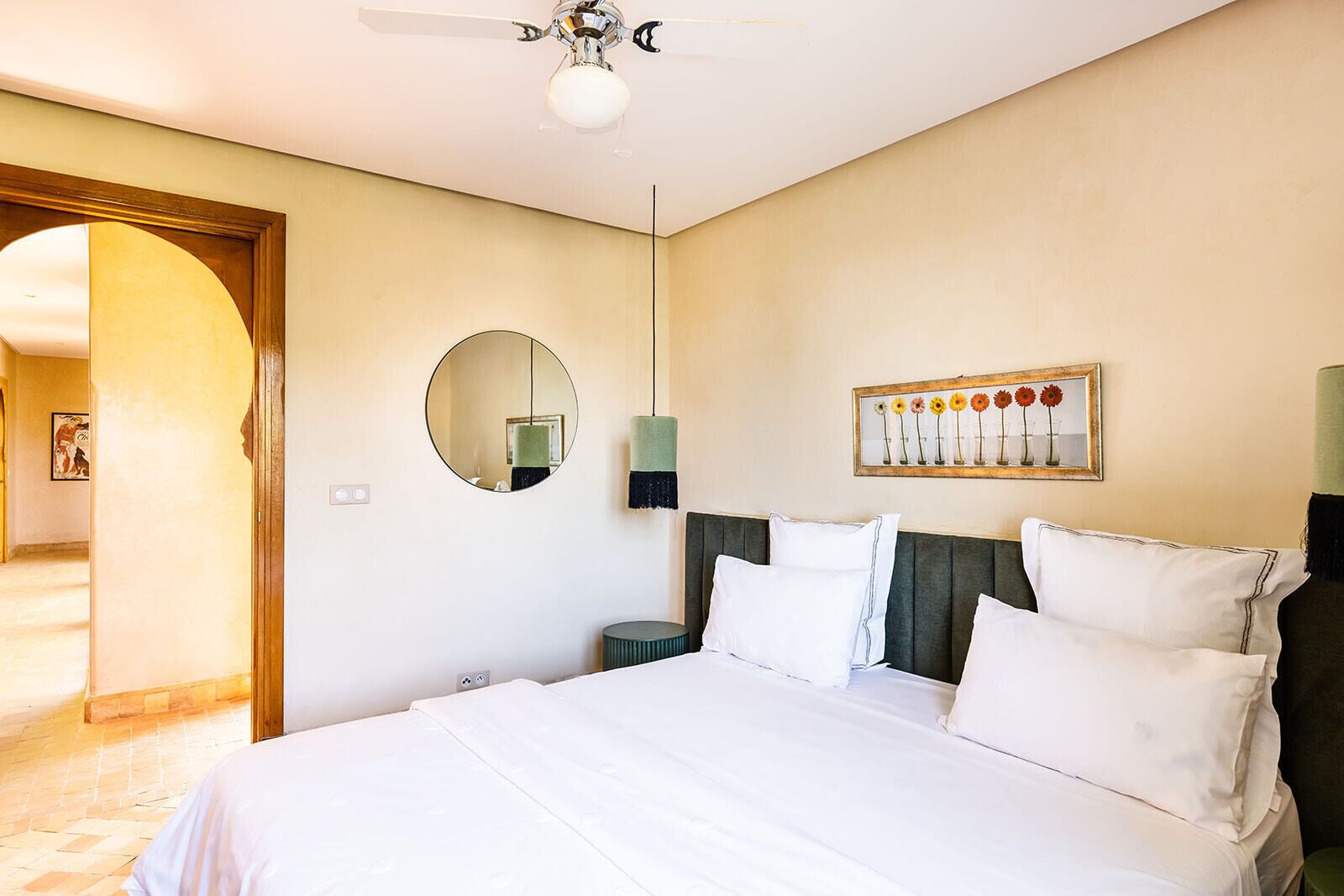 Une chambre à coucher avec un lit, un miroir rond, un tableau accroché au mur et un ventilateur de plafond.