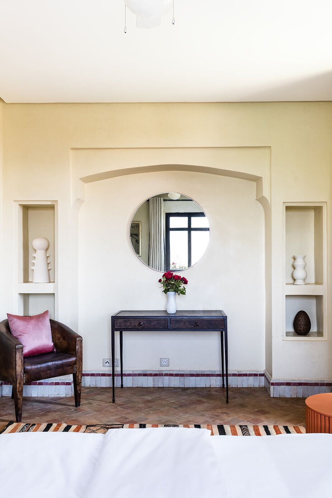 Una habitación con un espejo redondo, una consola de madera oscura con un jarrón de flores rojas, un sillón de cuero marrón con un cojín rosa y una alfombra estampada.