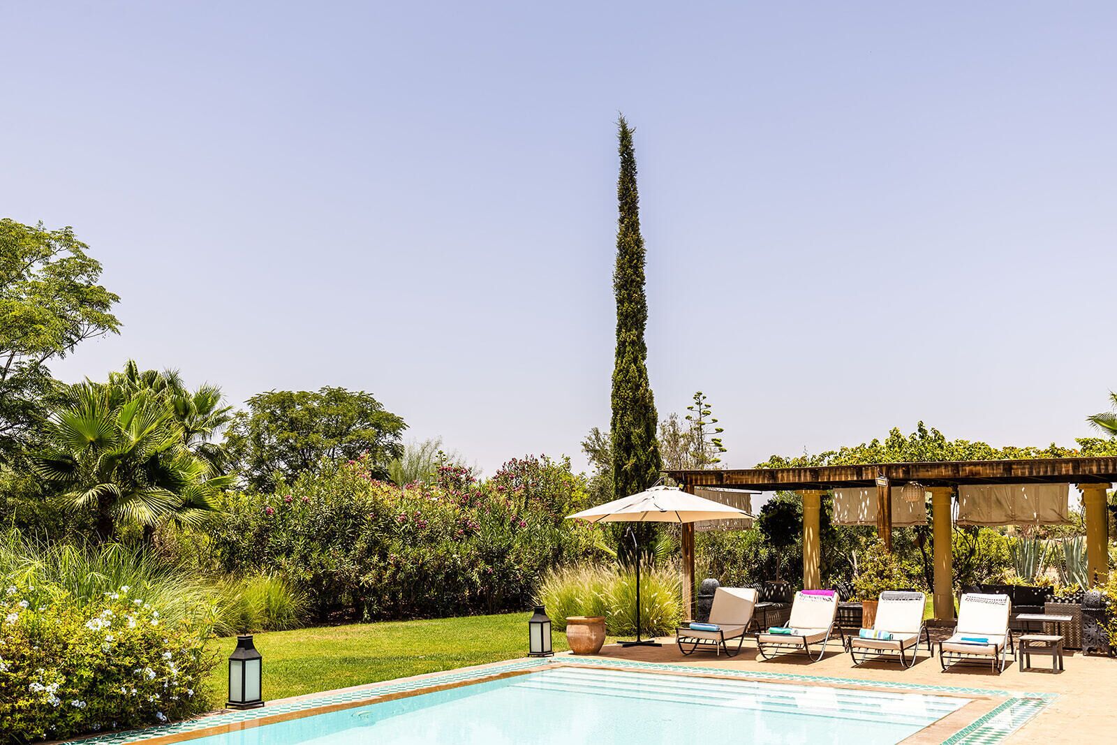 Un espace aménagé au bord de la piscine, avec des chaises longues, une pergola et un grand arbre.