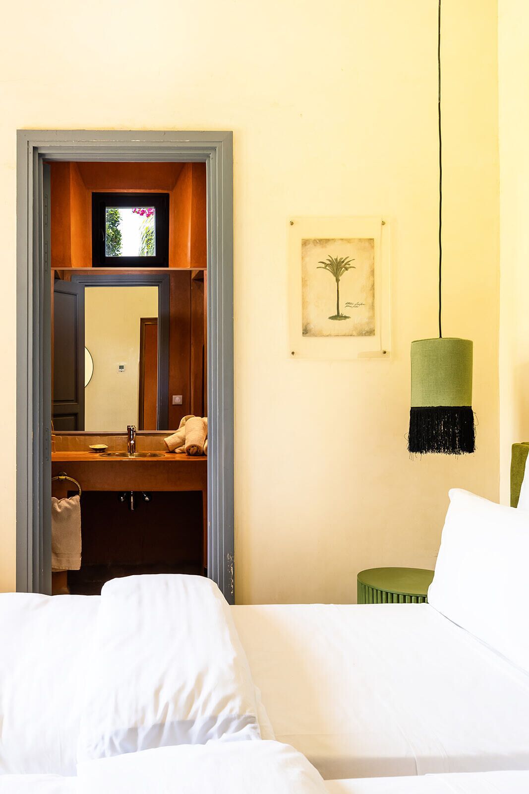 A hotel room with a bed, a green side table, a hanging lamp, a framed picture on the wall, and a bathroom visible through an open door.