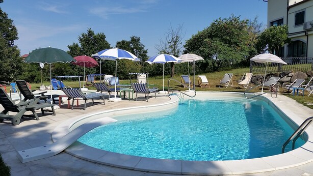 Una piscina con sillas de descanso y sombrillas, rodeada de vegetación y un edificio al fondo.