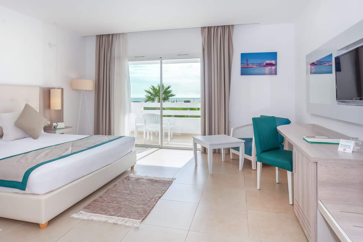 A hotel room with a bed, a desk, a chair, a TV, and a balcony with a view of the ocean.
