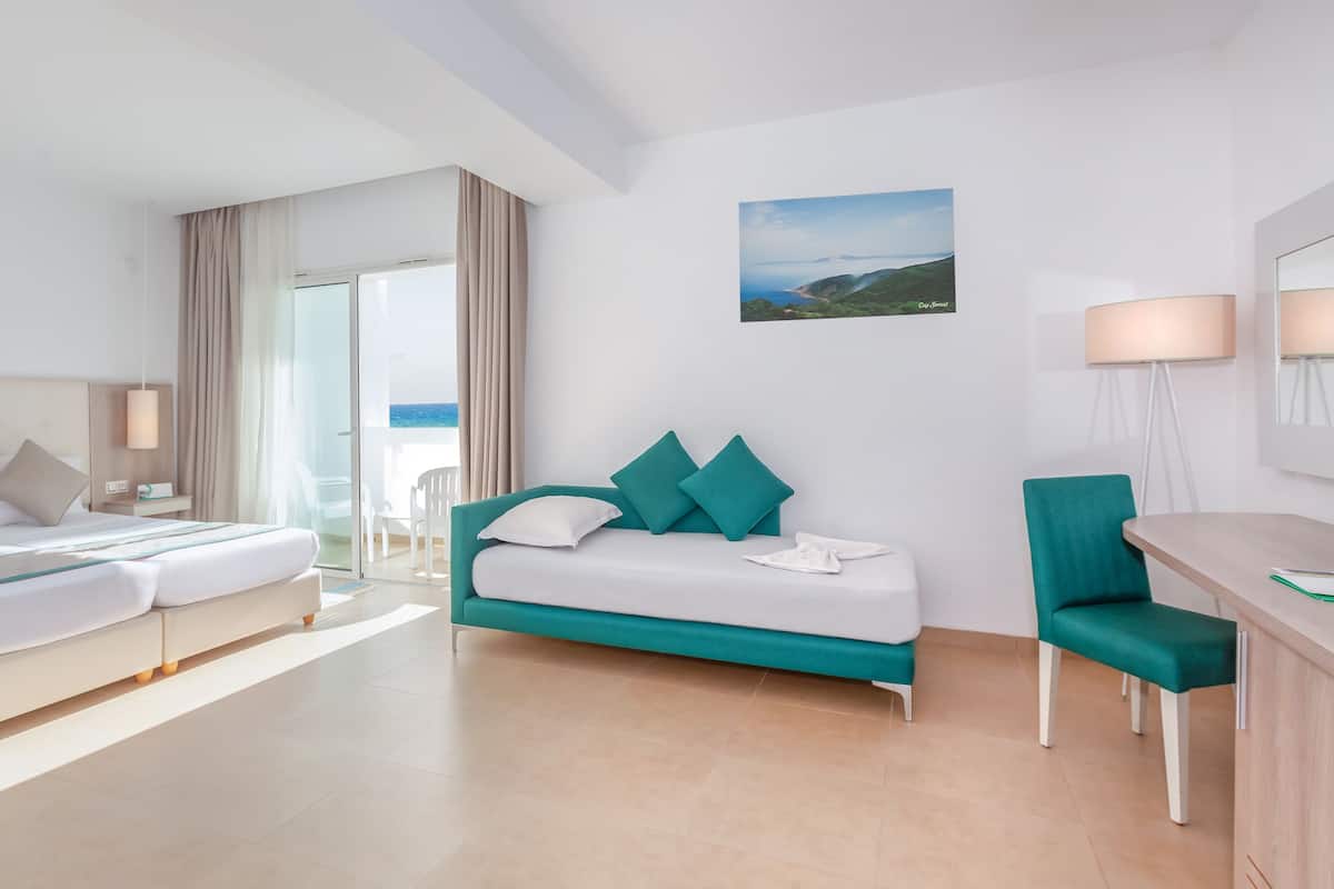 A modern hotel room with a bed, a sofa, a desk, and a view of the sea.