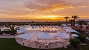 Un espace piscine aménagé dans un complexe hôtelier, avec des chaises longues et des parasols, entouré de palmiers et offrant une vue sur le coucher de soleil.