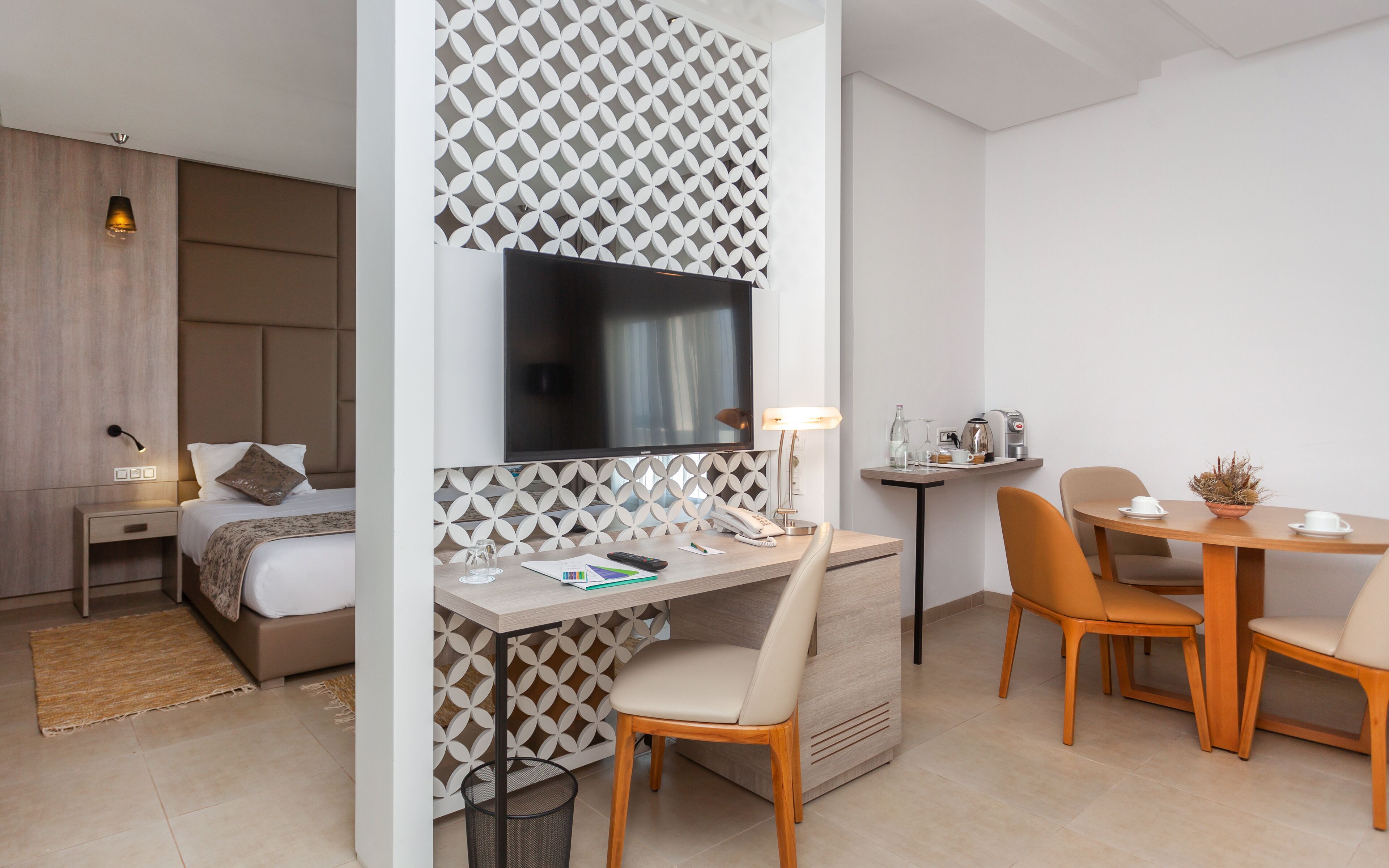 Una habitación de hotel moderna con una cama, un escritorio con silla, un televisor y un pequeño comedor con mesa y sillas.