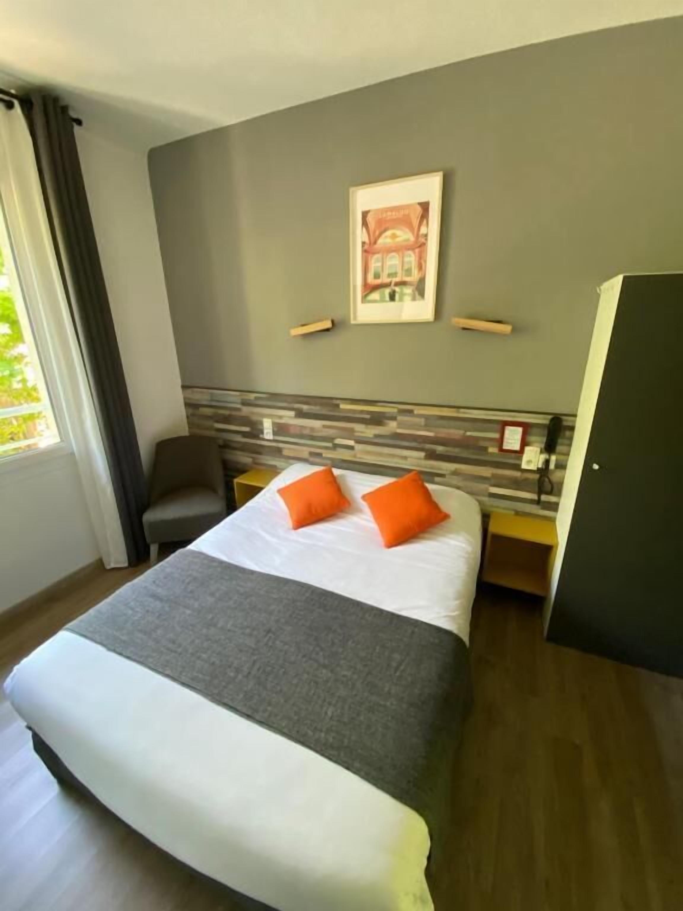 Una habitación de hotel con cama, una silla, escritorio, armario y ventana con cortinas.