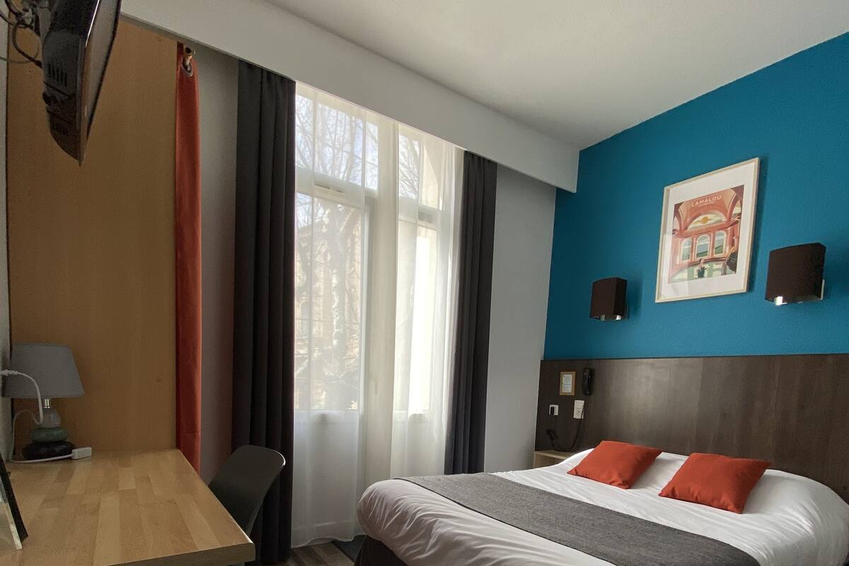 A hotel room with a bed, a desk, a chair, a TV, a window with curtains, and a wall with a framed picture.