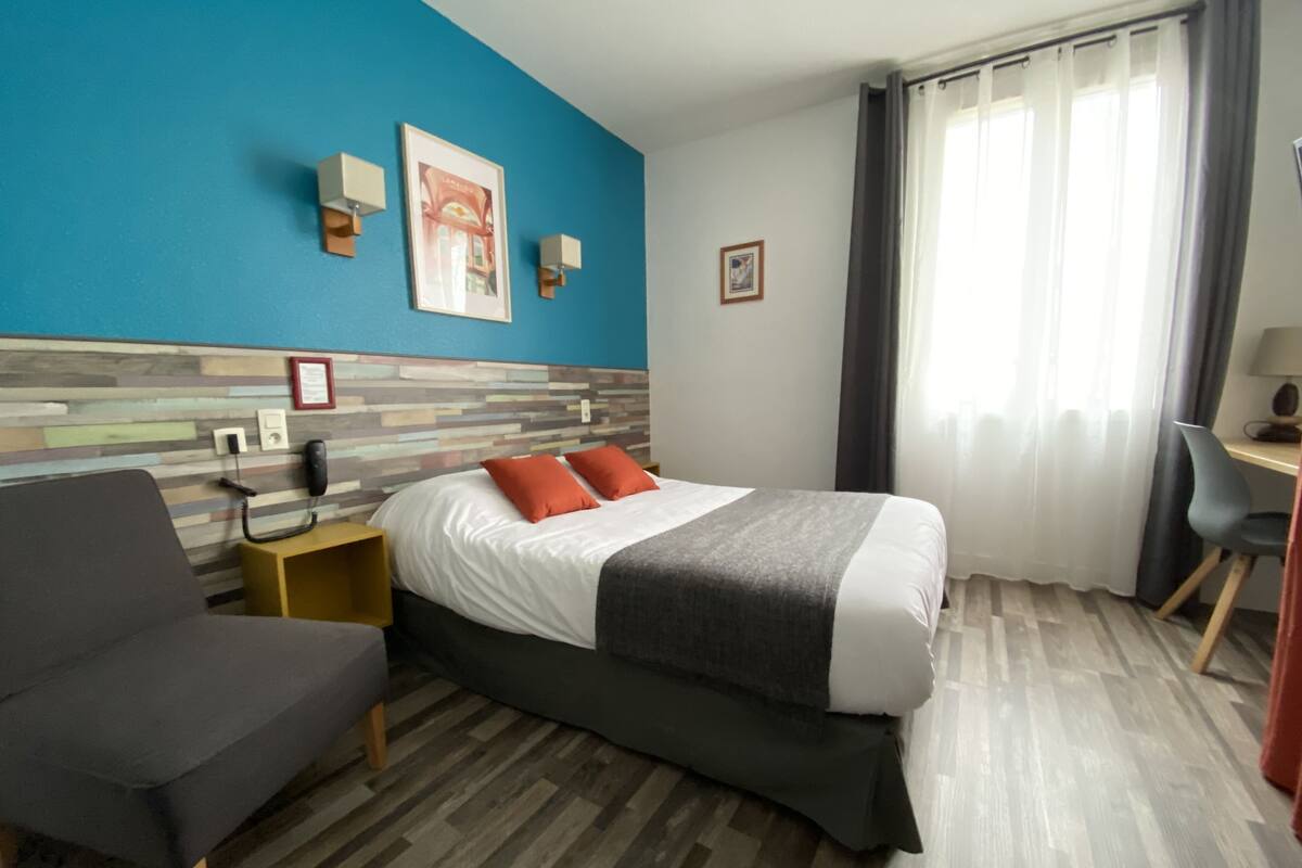 A hotel room with a bed, a desk, a chair, a phone, and a wall with a picture.