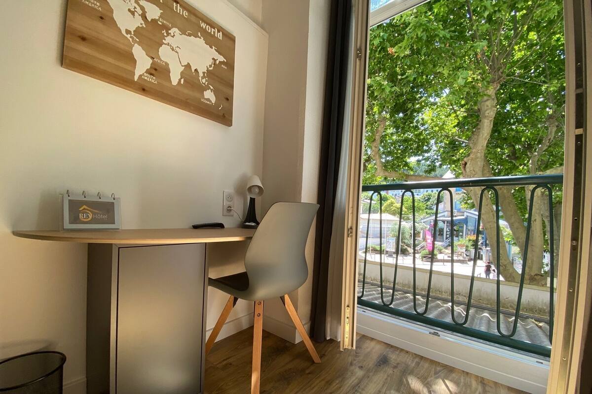 A hotel room with a desk, chair, and a view of the street through a window with a metal railing.