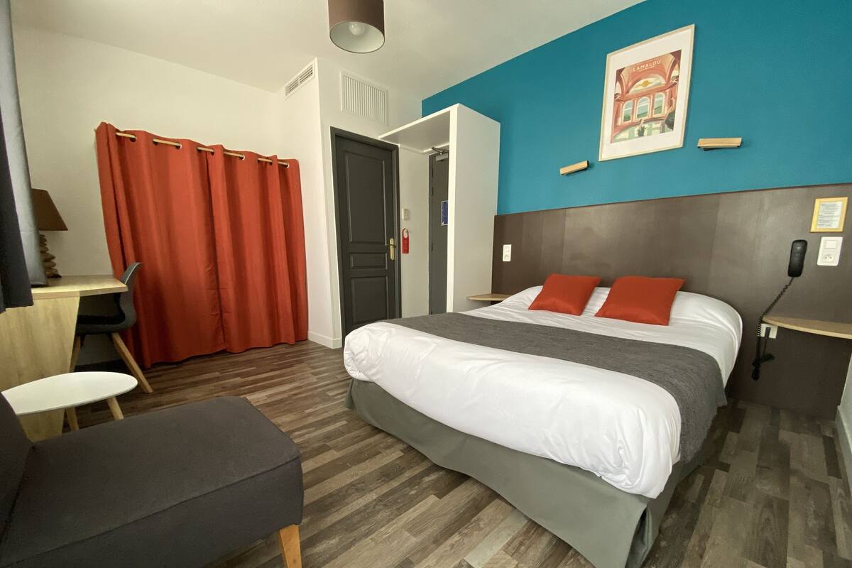 A hotel room with a bed, a desk, a chair, a desk lamp, a red curtain, and a framed picture on the wall.