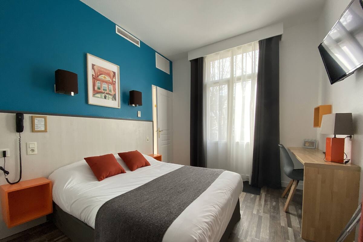 A hotel room with a bed, a desk, a chair, a TV, and a window with curtains.