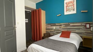 Habitación de hotel con cama, mesita de noche, silla, puerta y pared azul con un cuadro.