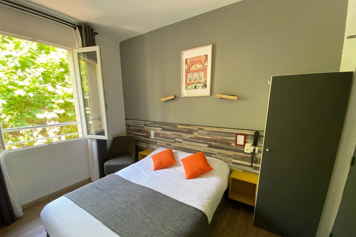 A hotel room with a bed, a chair, a desk, a wardrobe, a window with a view of trees, and a framed picture on the wall.
