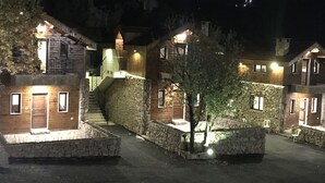 A nighttime view of a rustic lodge with stone walls, wooden cabins, and a central staircase leading to a second-floor balcony. - Villars (Mayrouba)
