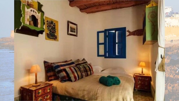 A bedroom with a bed, pillows, a nightstand, a lamp, and a window with blue shutters. - Riad Dar Latifa (Essaouira)