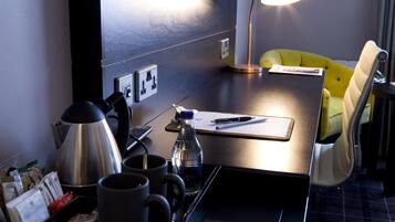 Habitación de hotel con escritorio, una silla amarilla, cafetera, botella de agua y lámpara.