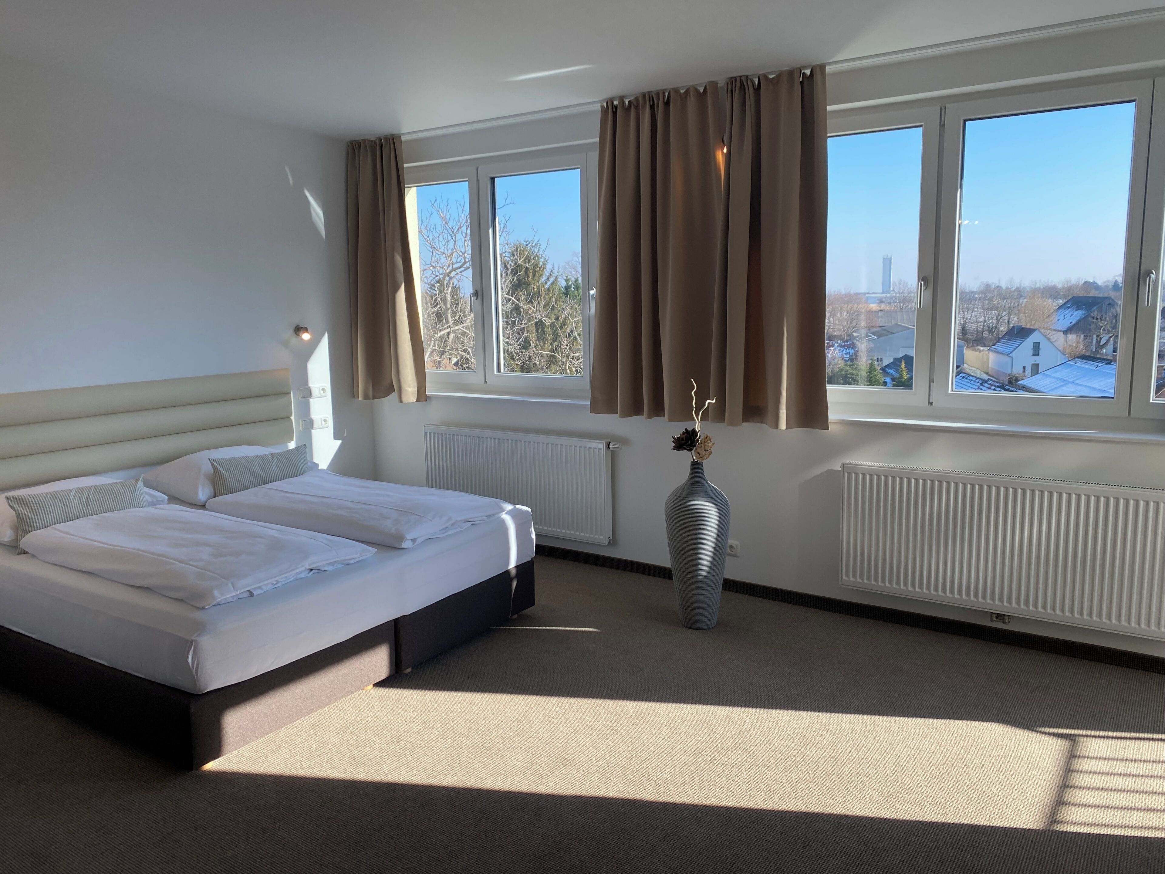 Habitación de hotel con cama doble, radiador, un florero con flores secas y vistas a edificios a través de amplios ventanales.