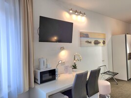 A modern hotel room with a flat-screen TV, a microwave, a small table with a coffee maker, a potted plant, and a seating area with chairs and a cushioned stool.