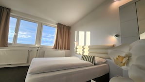 A hotel room with a large bed, a bedside table, a nightstand, a window with curtains, a radiator, and a wall-mounted light fixture. - Hotel Michael (Gerasdorf bei Wien)