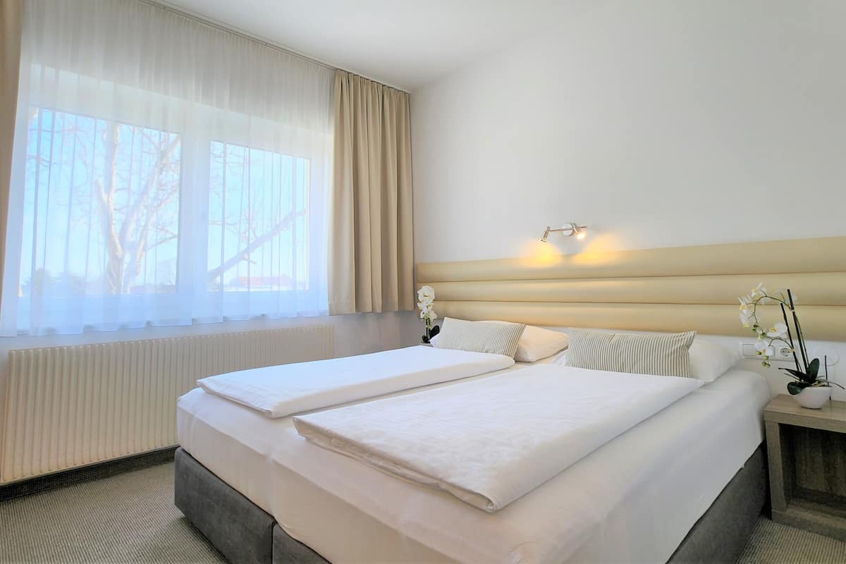 A hotel room with a large bed, a bedside table with a lamp, and a window with sheer curtains.