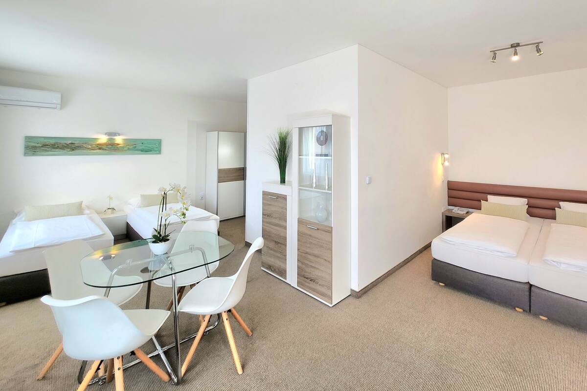 A modern hotel room with a glass dining table, white chairs, a bed with a brown headboard, and a small side table with a lamp.