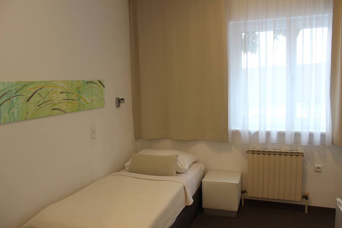 A small, single-bed room with a radiator, a window with sheer curtains, and a wall-mounted painting.