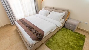 A neatly made bed with a patterned throw, a bedside table with drawers, and a green rug on a tiled floor. - PCG Gishushu Apartment (Kigali)