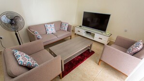 A living room with a sofa set, a coffee table, a TV on a stand, and a standing fan. - PCG Gishushu Apartment (Kigali)
