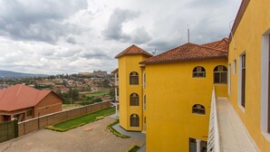 A yellow building with a red-tiled roof, a paved courtyard, and a view of a distant town. - PCG Gishushu Apartment (Kigali)