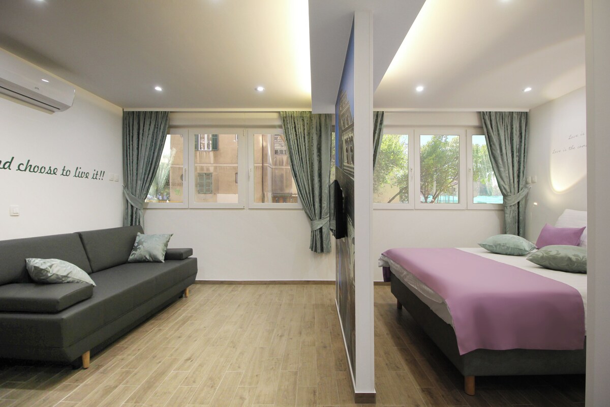 Ein modernes Zimmer mit einer Couch, einem Bett, einem Fernseher und großen Fenstern mit Vorhängen.