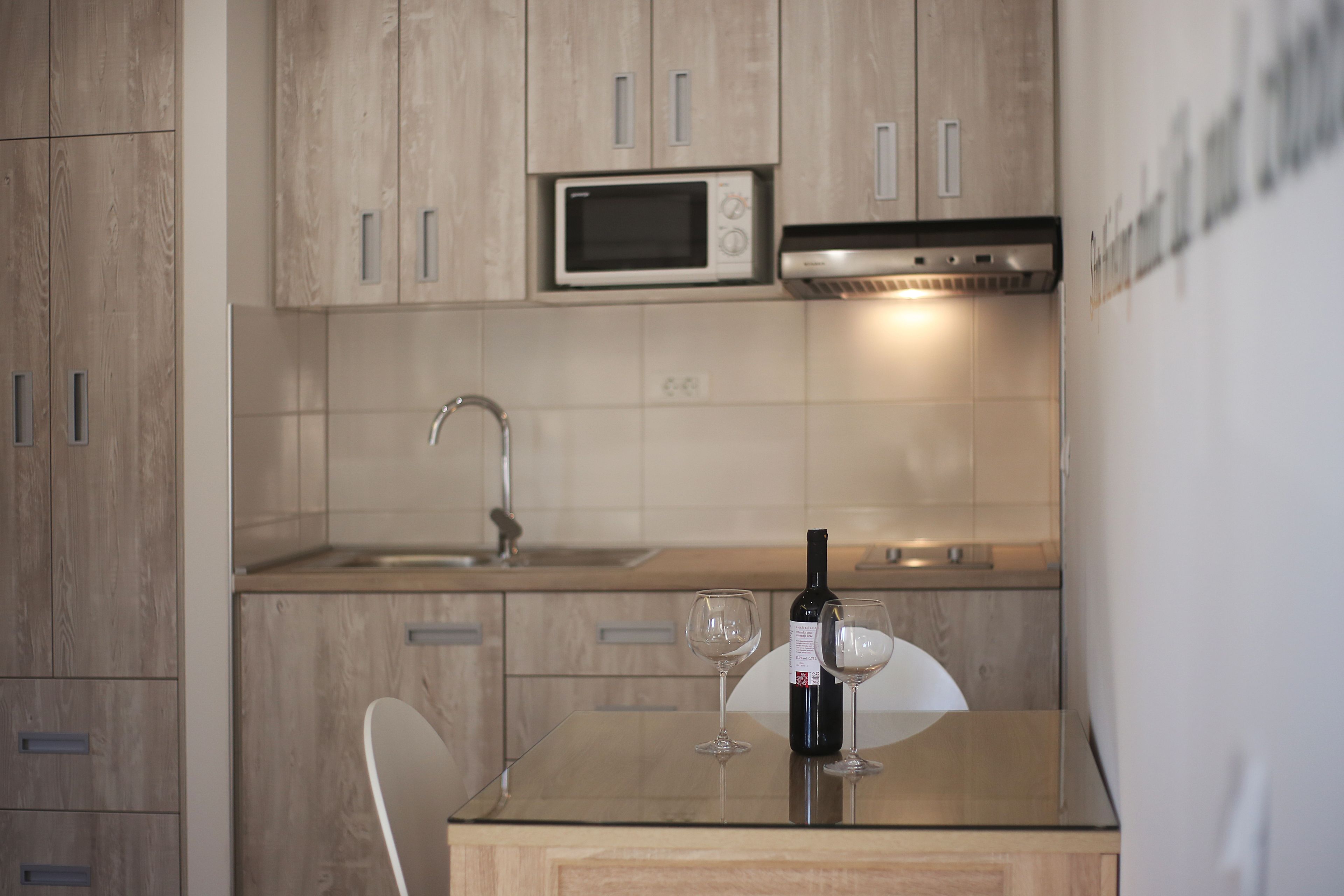 A kitchen with wooden cabinets, a microwave, a stainless steel range hood, a sink, and a glass table with a wine bottle and two wine glasses.