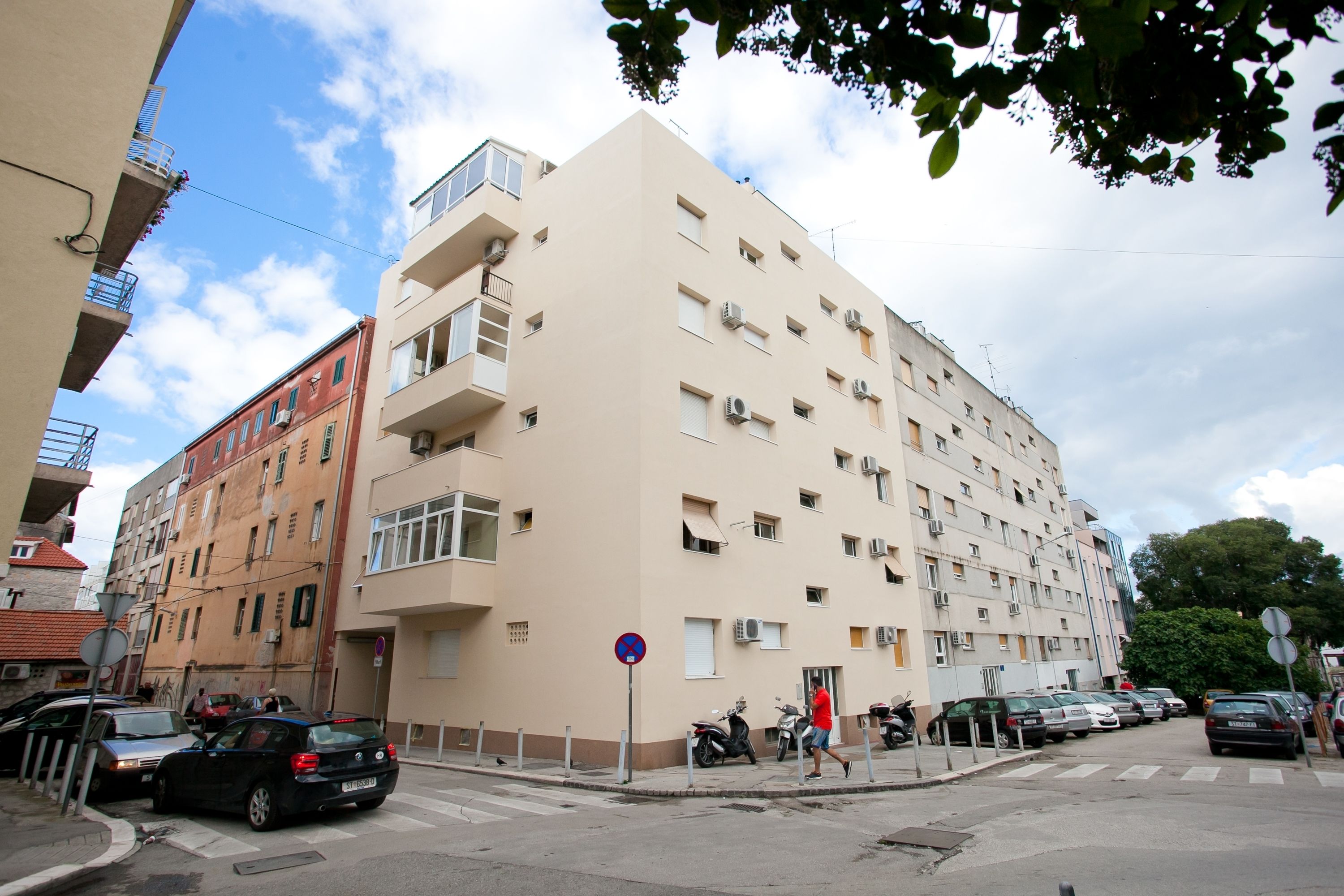 Un immeuble à plusieurs étages doté de balcons, avec des voitures garées et un passage piéton.