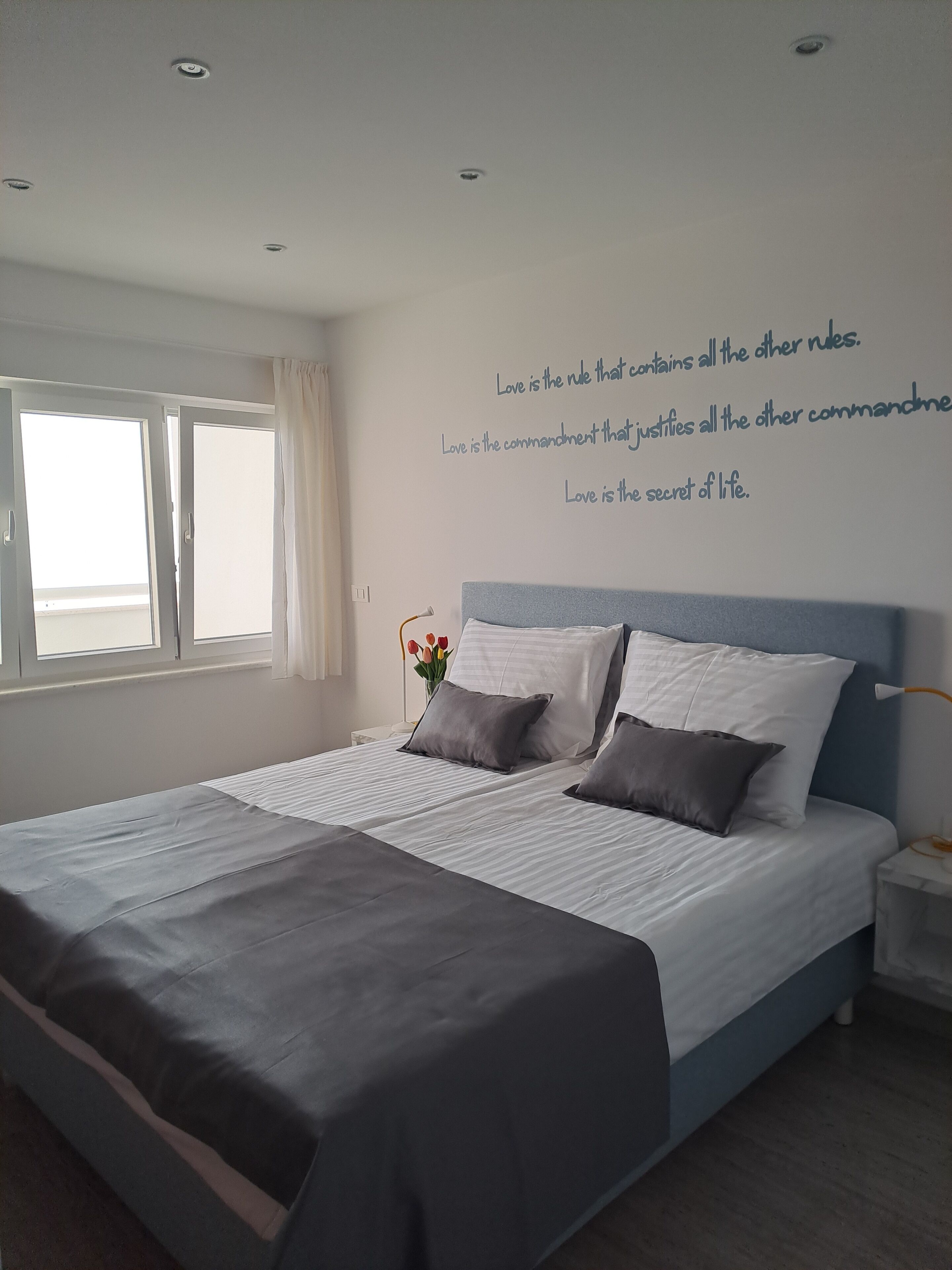 A bedroom with a large bed, a nightstand, a wall with inspirational quotes, and a window with curtains.