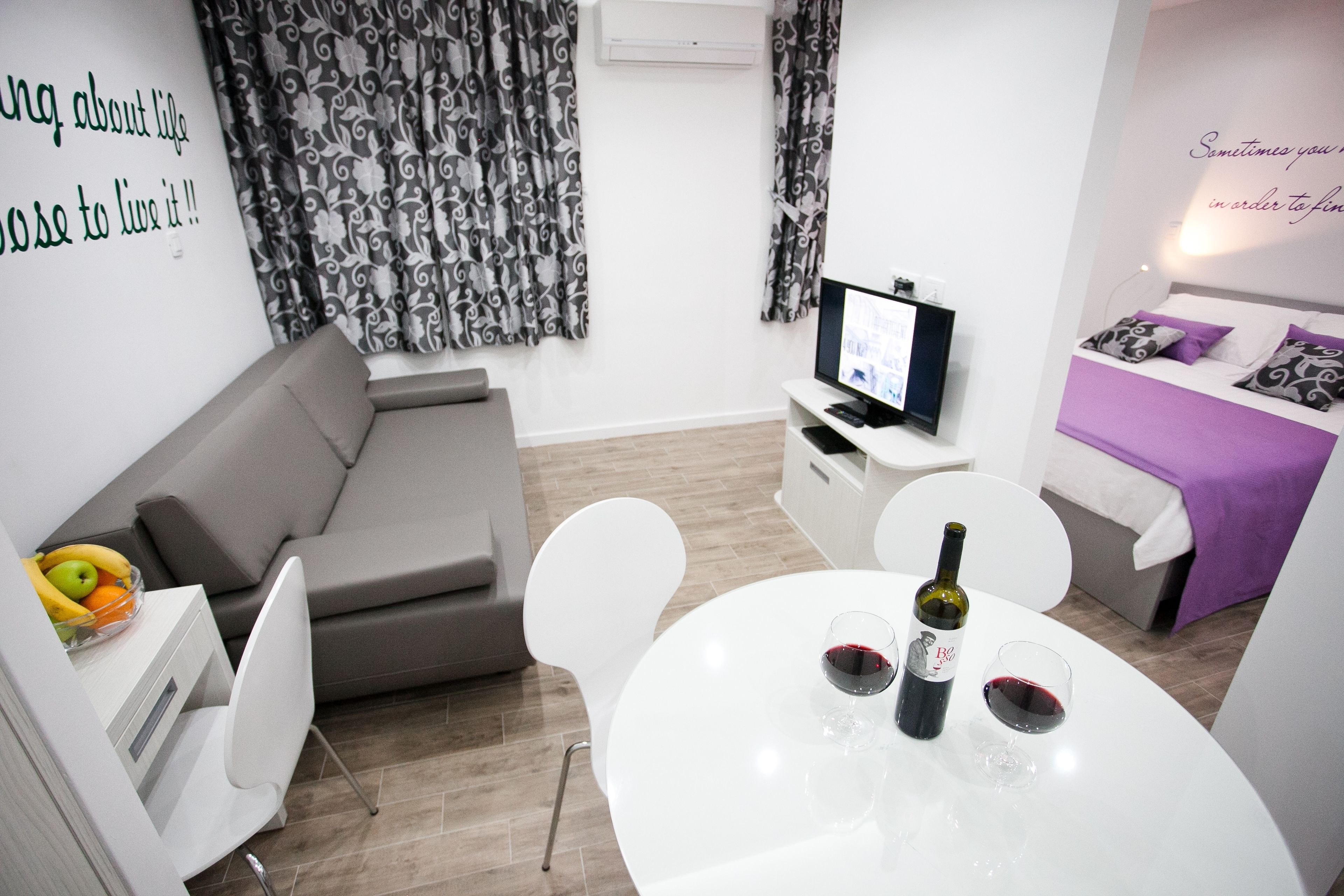 A modern living room with a grey sofa, a white dining table with two wine glasses and a bottle, a flat-screen TV on a white stand, and a bed with a purple bedspread.