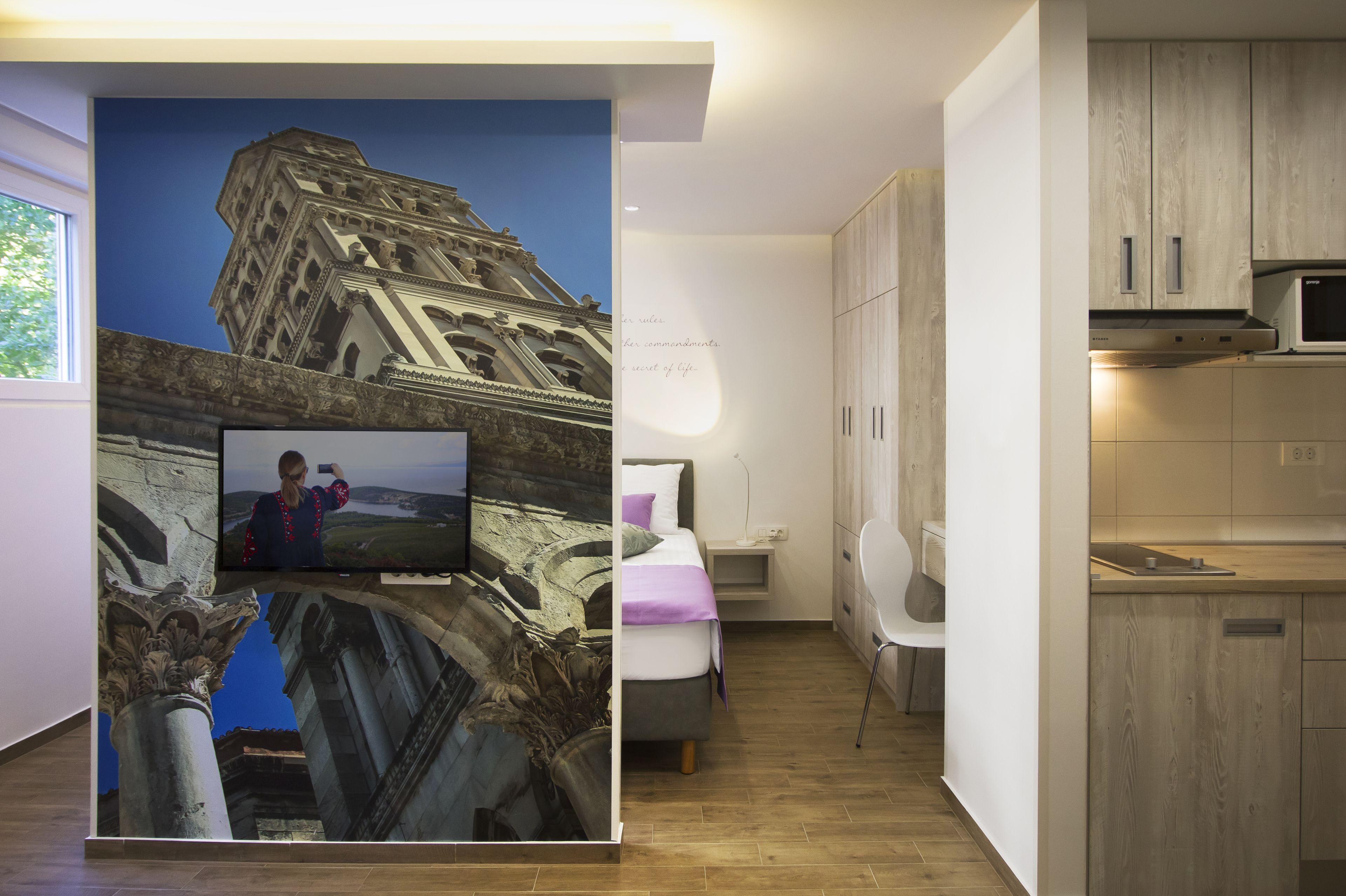 Habitación de hotel moderna con un gran mural de un edificio histórico, una cama con manta púrpura, un escritorio y una kitchenette.