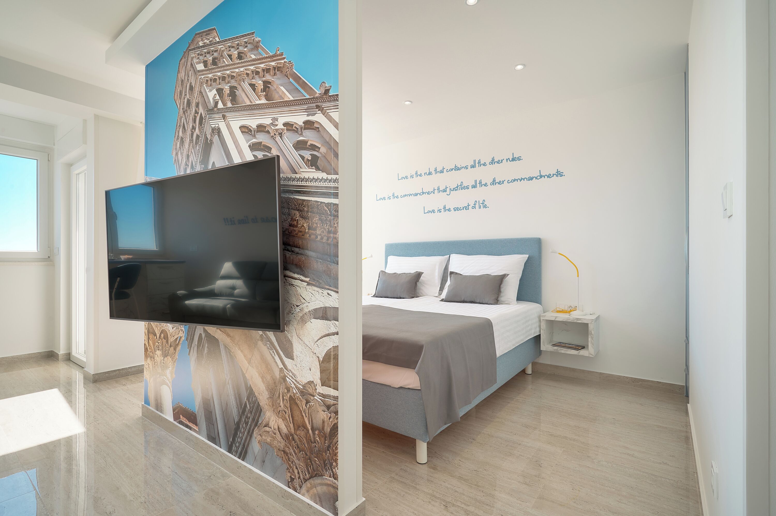 Una habitación de hotel moderna con una televisión de pantalla plana grande montada en la pared, una cama con cabecera azul y una mesita de noche pequeña con una lámpara.