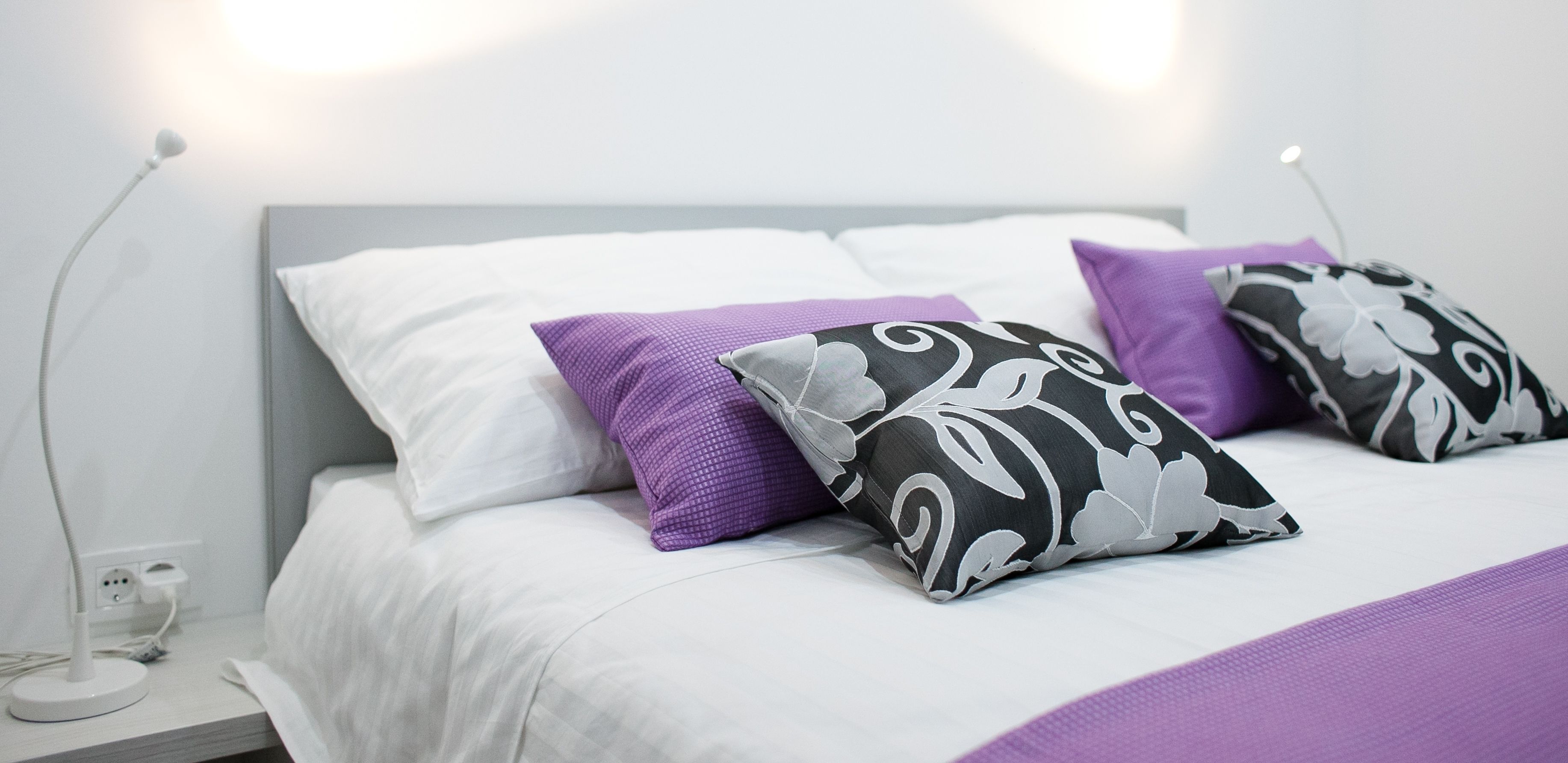 Una cama bien hecha con ropa de cama blanca y dos almohadas decorativas, una morada y otra con estampado blanco y negro.