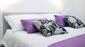 Una cama bien hecha con ropa de cama blanca y dos almohadas decorativas, una morada y otra con estampado blanco y negro.