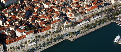 An aerial view of a coastal city with historic buildings, a waterfront promenade, and a ship docked at the port.