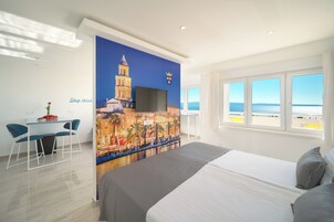 Ein Hotelzimmer mit einem Bett, einem Schreibtisch mit Stühlen, einem großen Fenster mit Meerblick und einem Wandbild einer Stadtlandschaft.