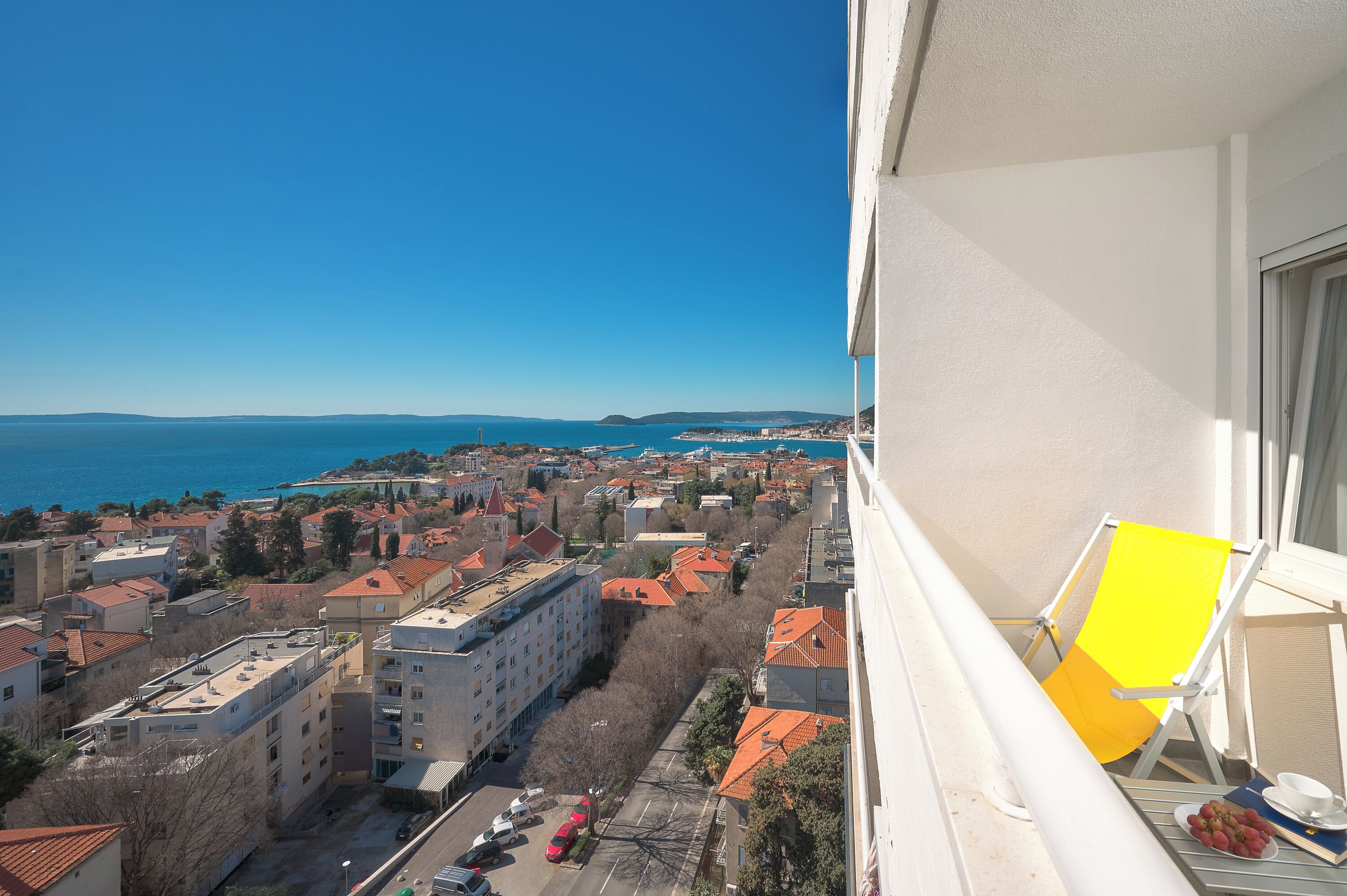 Un balcón con una silla amarilla, una mesa con una taza y uvas, y vistas a un pueblo costero con edificios y el mar.