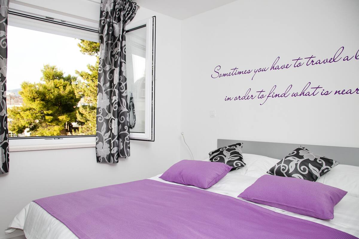Une chambre à coucher avec un lit, une literie violette, des rideaux à motifs et une citation peinte sur le mur.