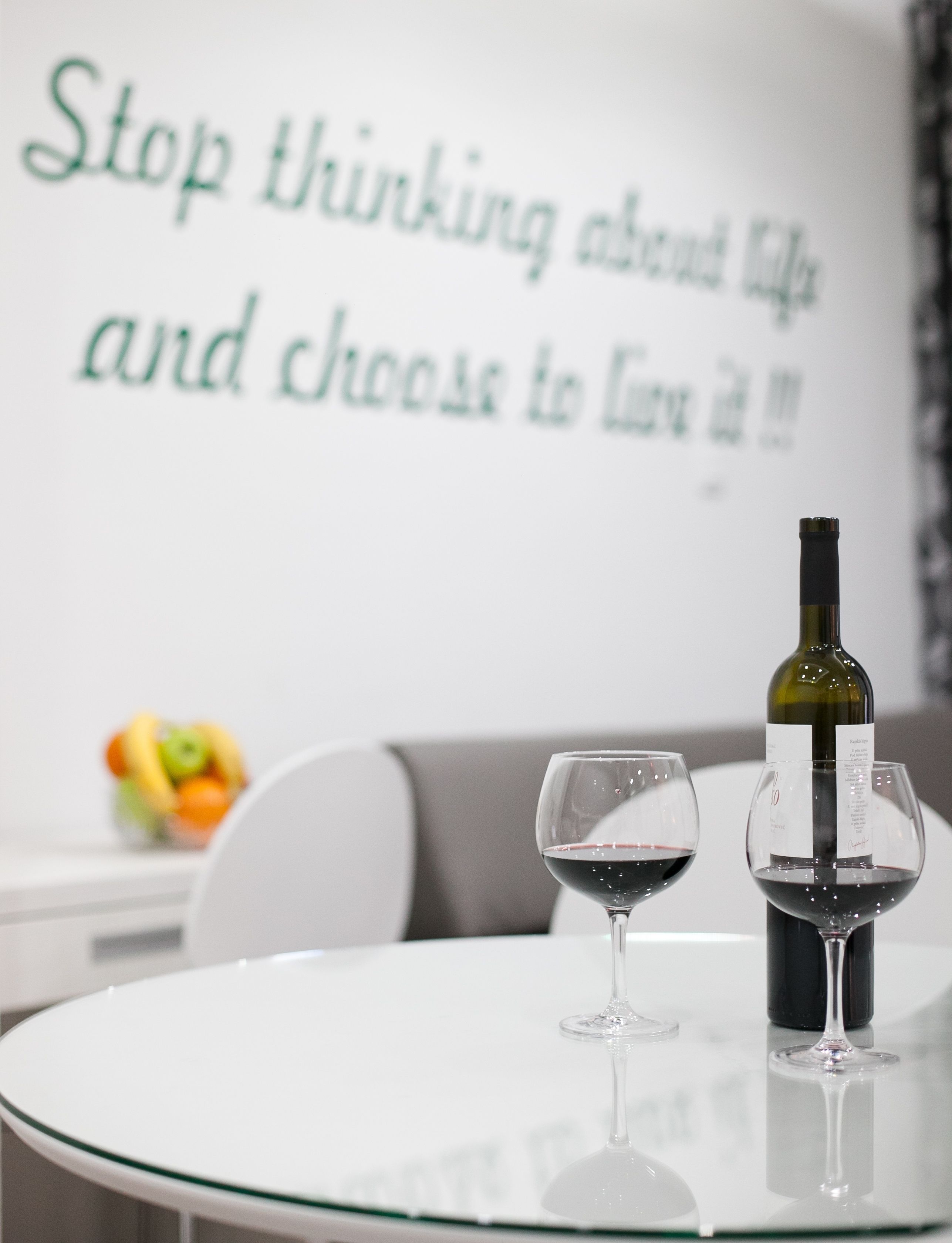 Een tafel met een wijnfles en twee glaasjes, een fruitschaal, en een inspirerende quote op de muur.