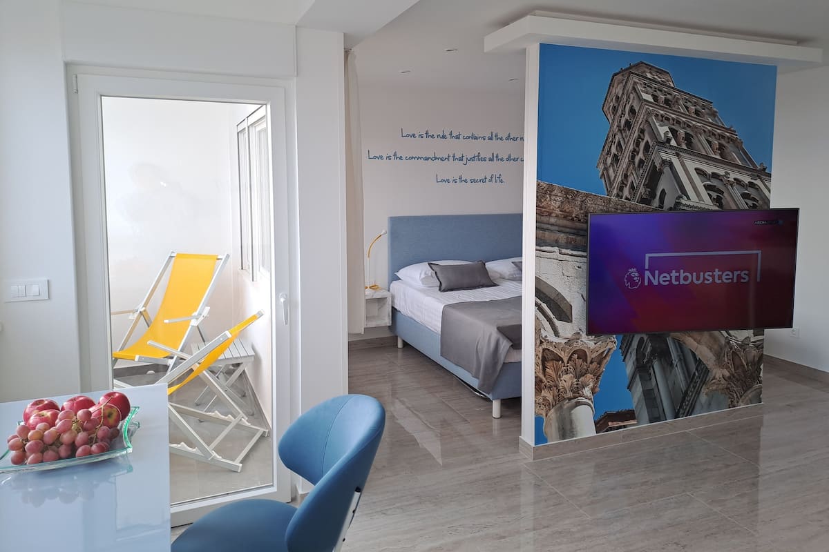 Une chambre d’hôtel avec un lit, un bureau et une chaise, une table avec des fruits, une fenêtre et un mur sur lequel est accrochée une télévision Netbusters.