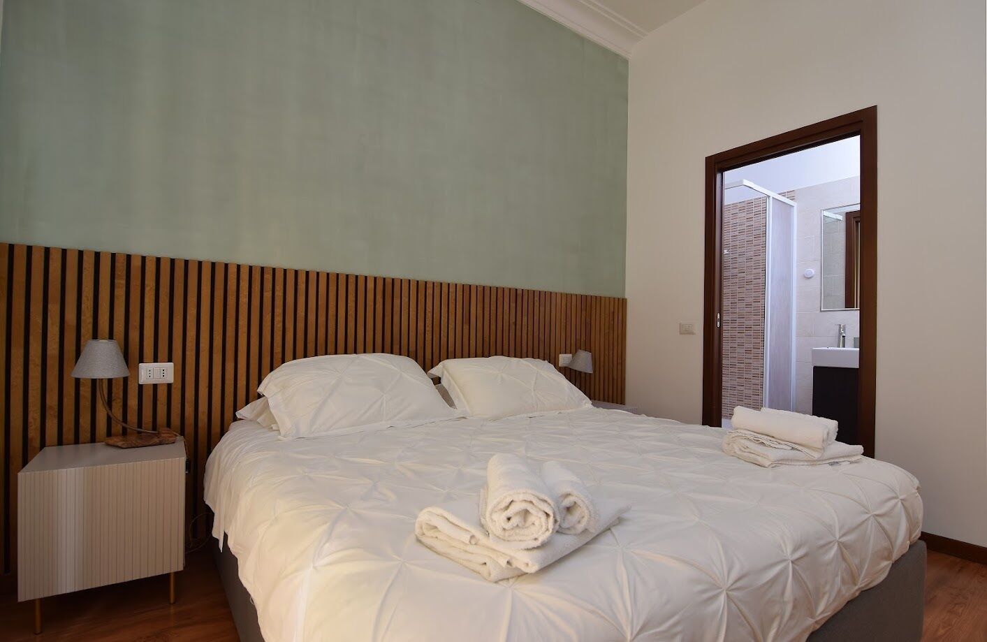 Una cama bien hecha con sábanas blancas y una toalla enrollada, una lámpara de noche y un cabecero de madera.