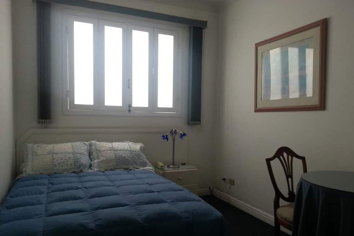 Ein Schlafzimmer mit Bett, Nachttisch, Stuhl, Tisch und einem Fenster mit Jalousien.